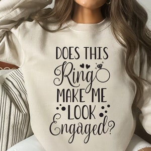 May include: A beige sweatshirt with the text "Does this ring make me look engaged?" in black lettering. The text includes a stylized ring with a diamond and two hearts.