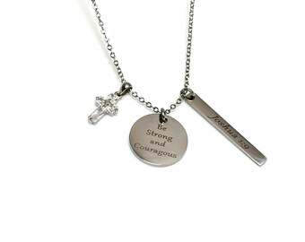 Puede incluir: Collar de plata con tres colgantes: una cruz con un cristal transparente, un colgante redondo con las palabras "Sé fuerte y valiente", y un colgante rectangular con las palabras "Josué 1:9".