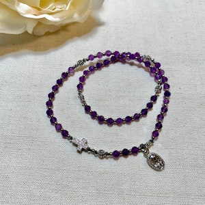 Puede incluir: Un rosario de cuentas moradas con un colgante de cruz plateada y un colgante ovalado. Las cuentas son de color morado intenso, y la cruz está incrustada con pequeñas piedras transparentes. El rosario se exhibe sobre una superficie blanca.