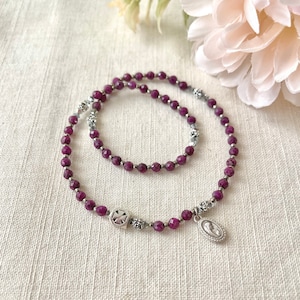 Puede incluir: Dos pulseras de piedras preciosas púrpuras con detalles plateados. Las pulseras presentan pequeñas flores plateadas y un dije plateado con una imagen religiosa.