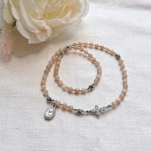 May include: A beaded rosary necklace featuring smooth, light peach-colored round beads interspersed with small silver-toned metal rose spacers. The necklace includes a silver-toned oval pendant depicting a figure and a small, clear crystal-studded cross.