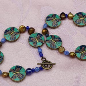 May include: A beaded bracelet featuring round, teal-colored charms with butterfly designs. The bracelet also includes blue and gold-colored beads, and a toggle clasp. The jewelry is set against a pink background.