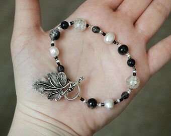 May include: A beaded bracelet featuring black, white, and clear beads, along with a silver leaf charm. The bracelet includes a toggle clasp and is displayed on a hand.