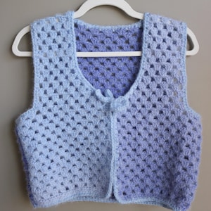 May include: A crocheted vest with a light blue to purple ombre color gradient. The vest has a V-neck and a tie closure at the front.