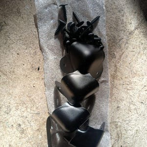 May include: A collection of black speaker corner protectors and screws. The corner protectors are L-shaped and appear to be made of metal. The screws are black and have a pointed end. The text "HEMP-O SPEAKERS JASON TICE" is visible.