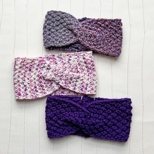 Könnte beinhalten: Drei handgestrickte Stirnbänder in Lila-, Lavendel- und Weißtönen. Die Stirnbänder sind aus dickem Garn gefertigt und haben ein verdrehtes Design. Die Farben sind eine Mischung aus einfarbigen und melierten Garnen, wodurch ein strukturierter Look entsteht.