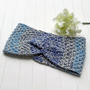 Könnte beinhalten: Ein handgefertigtes, blaues, graues und hellblaues gehäkeltes Stirnband. Das Stirnband ist aus weichem Garn gefertigt und hat ein verdrehtes Design. Eine weiße Blume befindet sich im Hintergrund.