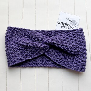 Könnte beinhalten: Lilafarbener Strick-Stirnband mit einem gedrehten Design. Das Stirnband ist mit einem strukturierten Stich gearbeitet und hat einen schlichten, modernen Stil.