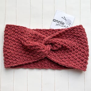 Könnte beinhalten: Ein rotes Strick Stirnband mit einem gedrehten Knotendesign. Das Stirnband ist aus einem dicken Garn gefertigt und hat eine weiche, kuschelige Textur.
