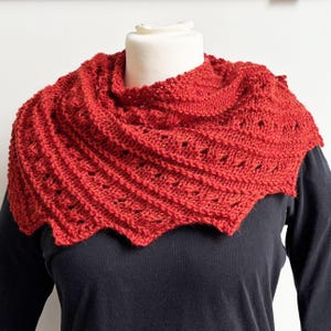 Könnte beinhalten: Ein roter gestrickter Schal mit einem Spitzenmuster. Der Schal ist über den Hals und die Schultern einer Schaufensterpuppe gelegt.