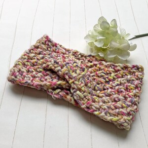 Könnte beinhalten: Ein handgestricktes Stirnband im Chunky-Strickstil. Das Stirnband hat eine Mischung aus Farben, darunter Rosa, Gelb, Lila und Creme. Das gemütliche Accessoire soll die Ohren warm halten. Eine weiße Blume befindet sich im Hintergrund.