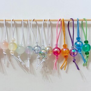 Könnte beinhalten: Eine Sammlung von bunten, schillernden und glitzernden Glasperlen an Schnüren mit Quasten. Die Perlen sind in verschiedenen Farbtönen von Rosa, Orange, Blau, Grün und Weiß.