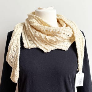 Könnte beinhalten: Cremefarbener Strickschal mit strukturiertem Design, drapiert auf einer Schaufensterpuppe. Der Schal hat eine dreieckige Form und komplizierte Strickmuster. Die weiche, neutrale Farbe und die gemütliche Textur suggerieren Wärme und Komfort. Ein Etikett ist an einem Ende befestigt.