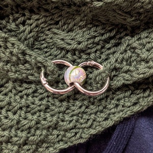 Könnte beinhalten: Nahaufnahme einer grünen Strickmütze mit einem silbernen Metallverschluss und einer weißen und grünen Glasperle.