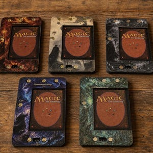 May include: Five custom card holders with the Magic: The Gathering logo and card back visible. The holders have unique designs in shades of brown, gray, blue, and green, with metallic accents. The cards are displayed on a wooden surface.