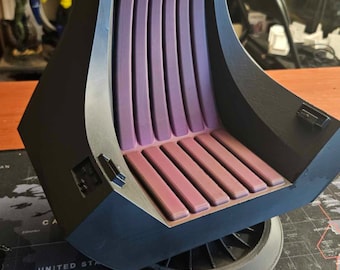 May include: A black and purple 3D-printed chair model with a detailed design. The chair features a curved back with vertical purple supports and a seat with horizontal red-brown slats. The model sits on a black base, placed on a world map.