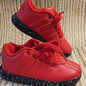 May include: A pair of red sneakers with white stitching and laces. The soles are black with white speckles. The shoes are displayed on a textured, neutral-toned surface.