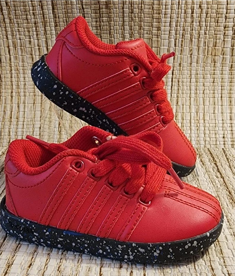 May include: A pair of red sneakers with white stitching and laces. The soles are black with white speckles. The shoes are displayed on a textured, neutral-toned surface.