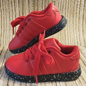 May include: A pair of red toddler sneakers with matching red laces. The shoes have a black sole speckled with white and blue flecks. The sneakers are displayed on a woven, light brown surface.