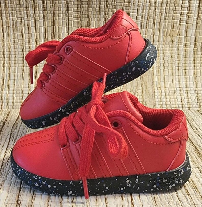 May include: A pair of red toddler sneakers with matching red laces. The shoes have a black sole speckled with white and blue flecks. The sneakers are displayed on a woven, light brown surface.