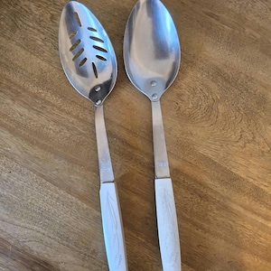 May include: Two stainless steel kitchen spoons with white handles. One spoon is solid, and the other has a slotted design. The handles have a decorative etched pattern. The spoons are resting on a wooden surface.