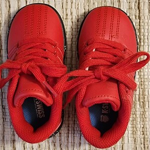 May include: Pair of bright red K-Swiss sneakers for toddlers. The shoes have red laces, a red upper, and a black sole. The K-Swiss logo is visible on the tongue and insole. The shoes are new and unworn.