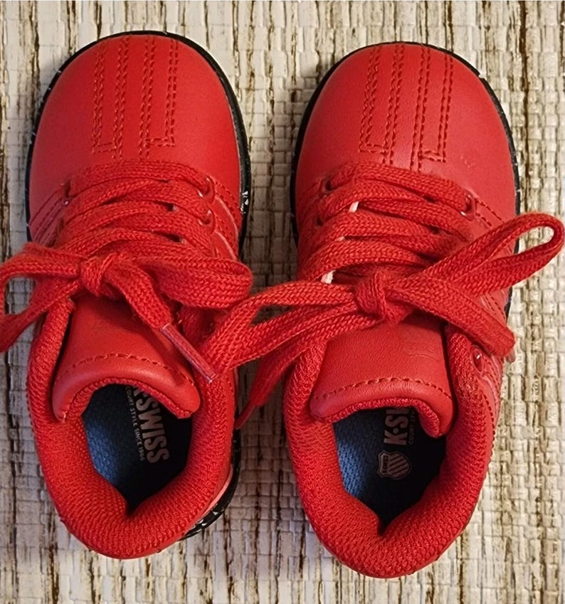 May include: Pair of bright red K-Swiss sneakers for toddlers. The shoes have red laces, a red upper, and a black sole. The K-Swiss logo is visible on the tongue and insole. The shoes are new and unworn.