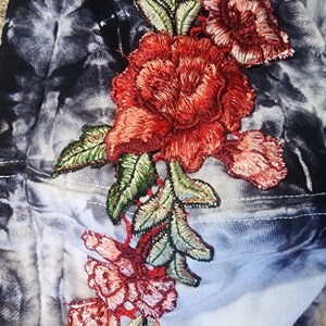May include: A tie-dye hoodie with a large embroidered floral design. The flowers are red and pink with green leaves. The background is a mix of blue, white, and black.