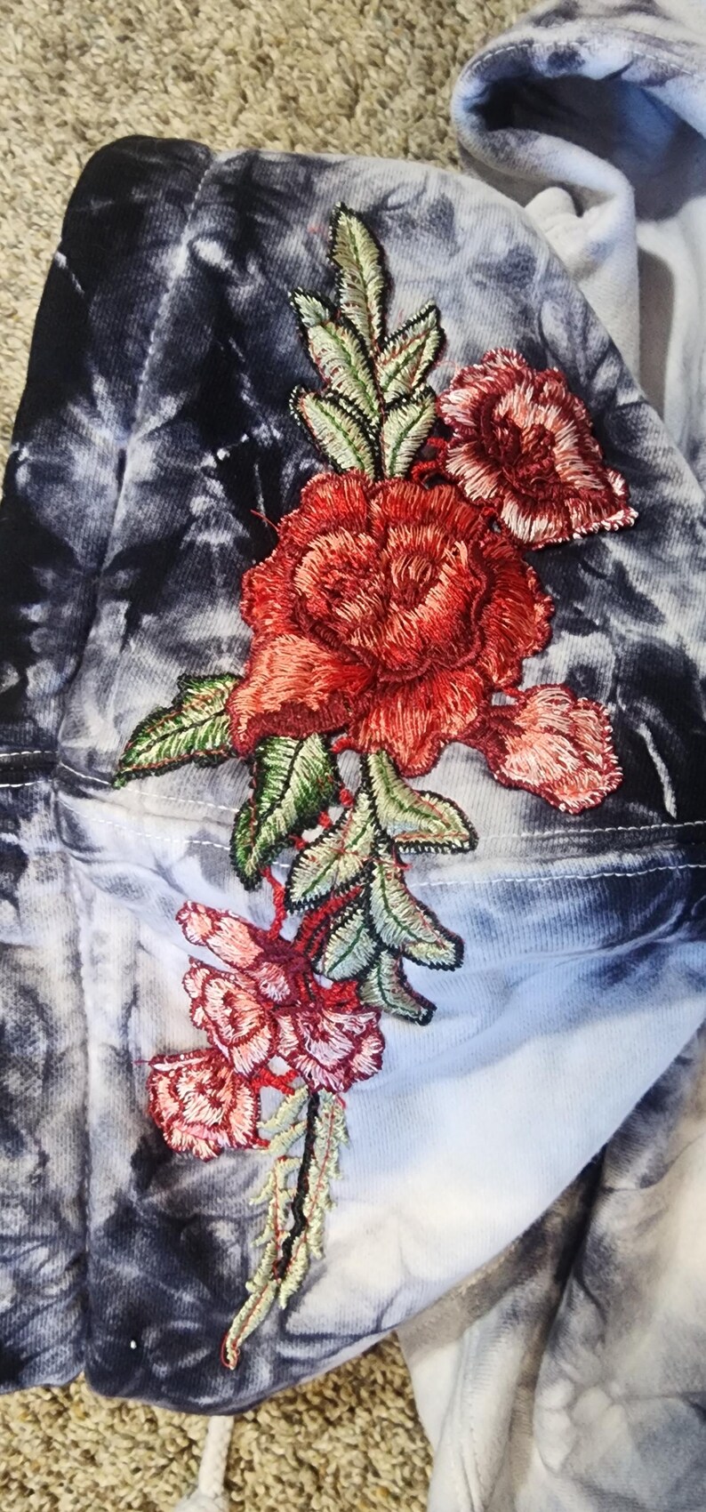 May include: A tie-dye hoodie with a large embroidered floral design. The flowers are red and pink with green leaves. The background is a mix of blue, white, and black.