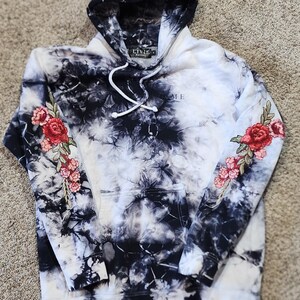 May include: A black and white tie-dye hoodie with a floral design on the hood and sleeves. The hoodie has a front pocket and a drawstring. The word "ME" is visible on the front of the hoodie.