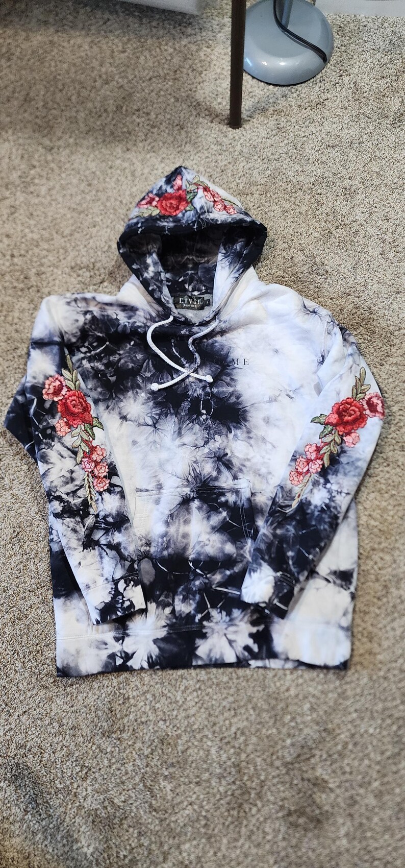 May include: A black and white tie-dye hoodie with a floral design on the hood and sleeves. The hoodie has a front pocket and a drawstring. The word "ME" is visible on the front of the hoodie.
