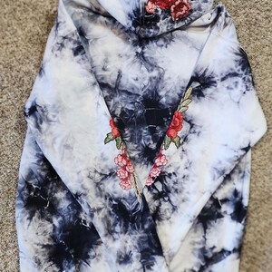 May include: A tie-dye hoodie in black and white with embroidered red roses and pink flowers on the hood and sleeves. The hoodie has a drawstring and a ribbed hem and cuffs. The overall style is casual and artistic.