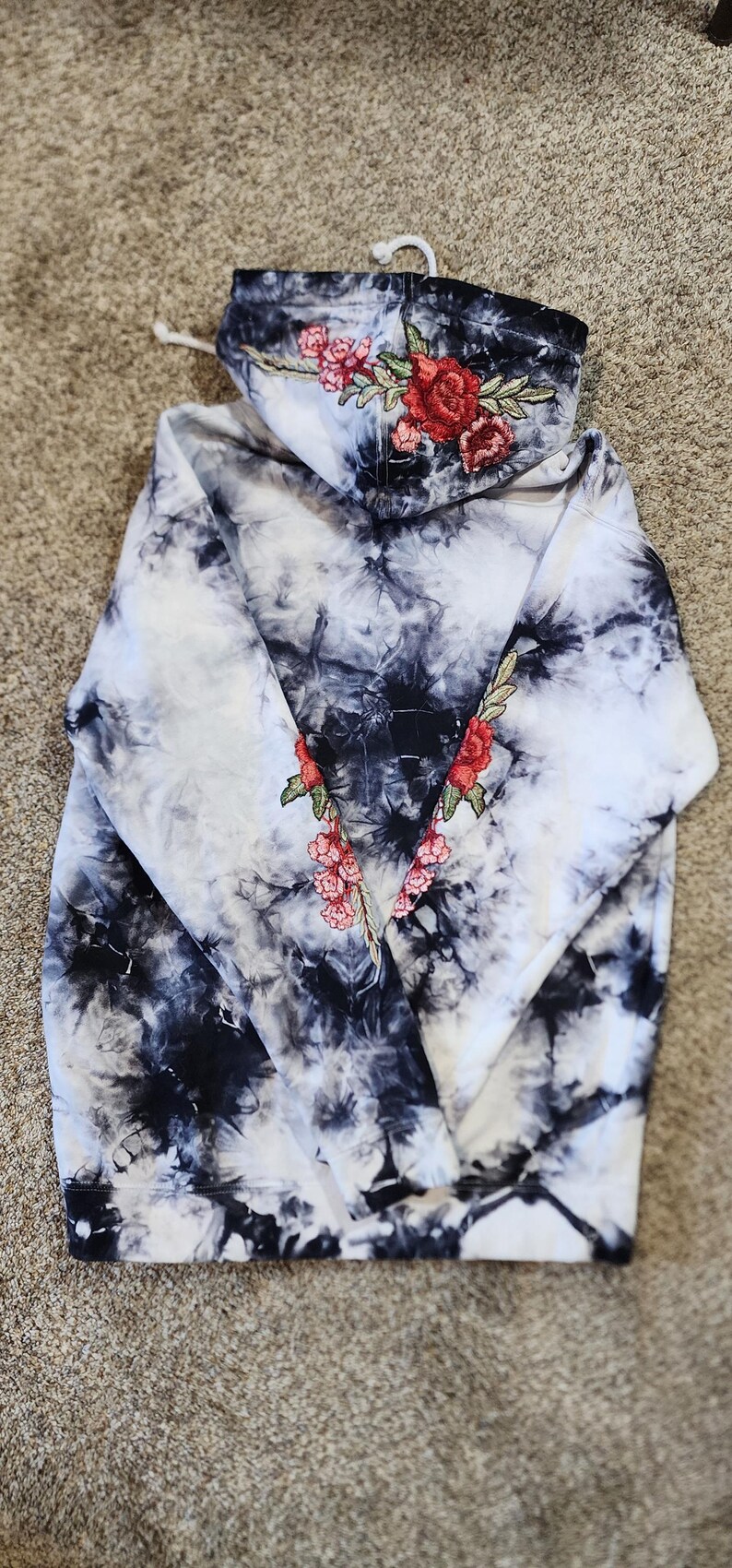May include: A tie-dye hoodie in black and white with embroidered red roses and pink flowers on the hood and sleeves. The hoodie has a drawstring and a ribbed hem and cuffs. The overall style is casual and artistic.