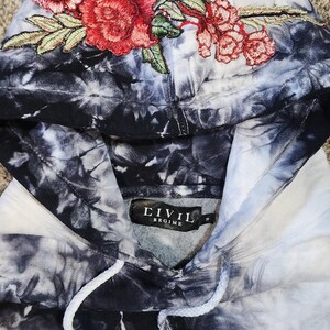 May include: A tie-dye hoodie in shades of black, white, and gray, featuring an embroidered floral design in red and green on the hood. The hoodie has a drawstring and a label that reads "CIVIL REGIME".