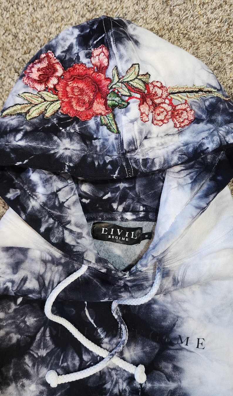 May include: A tie-dye hoodie in shades of black, white, and gray, featuring an embroidered floral design in red and green on the hood. The hoodie has a drawstring and a label that reads "CIVIL REGIME".