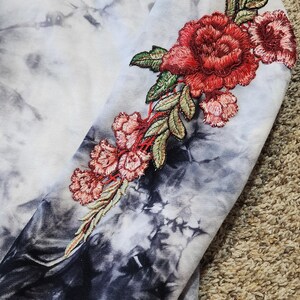 May include: A tie-dye hoodie with a floral embroidery design on the sleeve. The embroidery features red and pink roses with green leaves. The hoodie has a black and white tie-dye pattern.