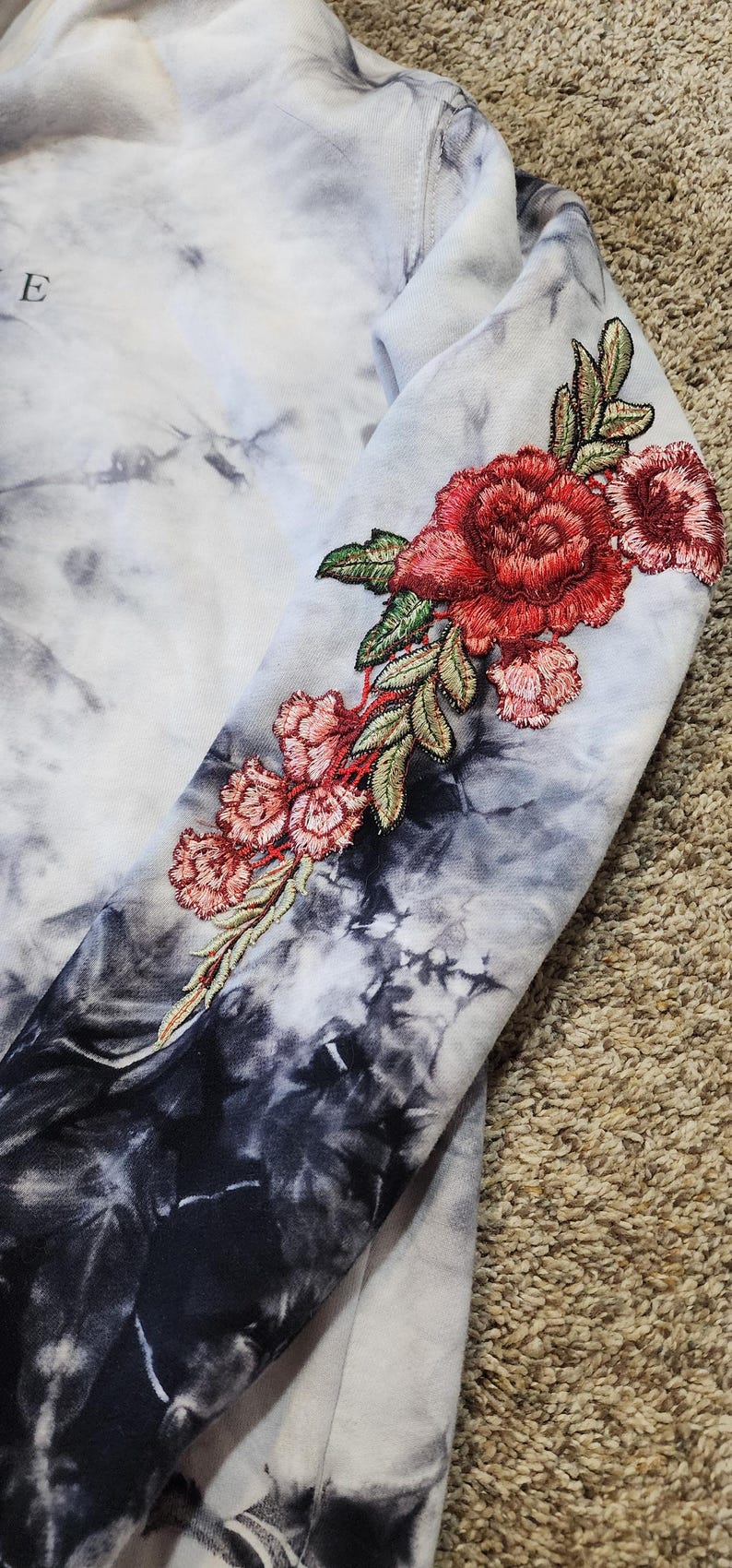 May include: A tie-dye hoodie with a floral embroidery design on the sleeve. The embroidery features red and pink roses with green leaves. The hoodie has a black and white tie-dye pattern.