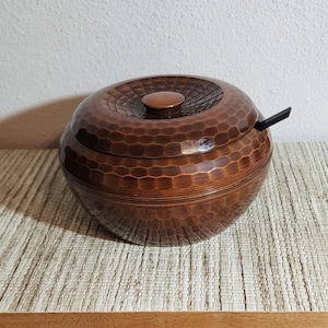 May include: A handcrafted, round wooden bowl with a lid and a small handle. The bowl is a rich brown color with a textured, hammered surface. The lid has a small, round knob. The bowl sits on a woven mat.