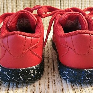 May include: A pair of red sneakers with matching laces. The shoes have a black sole speckled with white and blue accents. The back of the shoes are visible, showcasing the padded collar and stitching details.