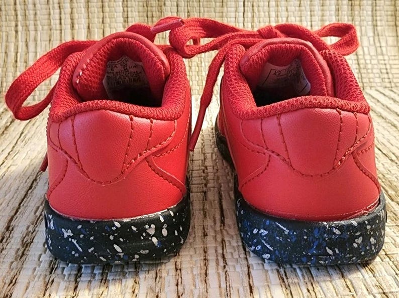 May include: A pair of red sneakers with matching laces. The shoes have a black sole speckled with white and blue accents. The back of the shoes are visible, showcasing the padded collar and stitching details.