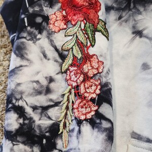 May include: A tie-dye hoodie in black, white, and gray, with an embroidered floral design in red, pink, and green. The floral design is on the sleeve of the hoodie.