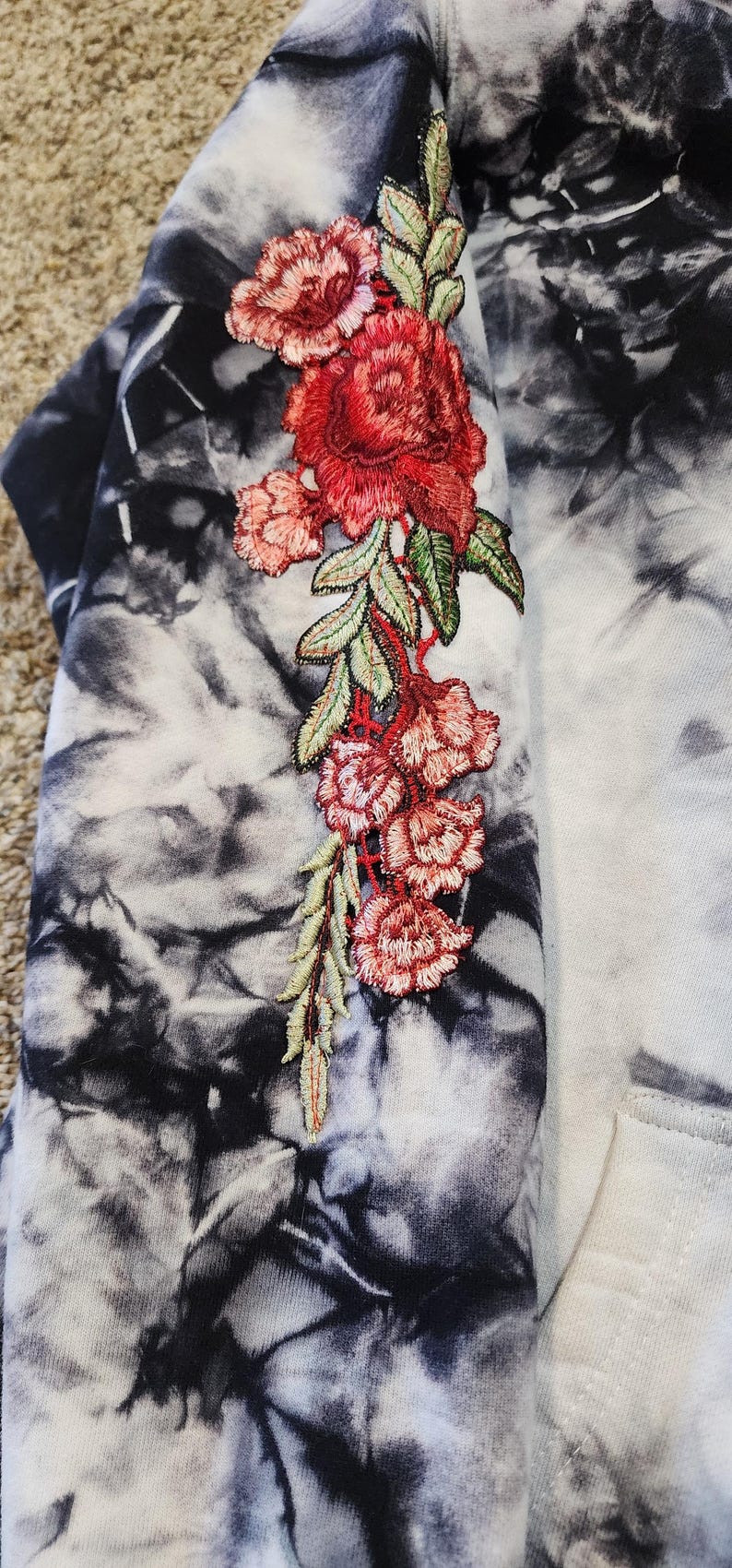 May include: A tie-dye hoodie in black, white, and gray, with an embroidered floral design in red, pink, and green. The floral design is on the sleeve of the hoodie.