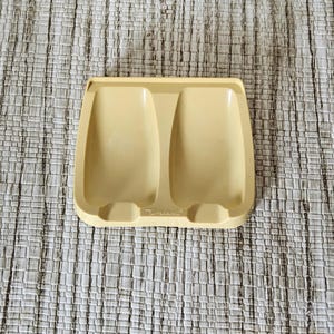 May include: A vintage, pale yellow Tupperware spoon rest. The rectangular dish has two spoon-shaped indentations and a raised center section. The word "Tupperware" is embossed on the front. The spoon rest is made of durable plastic.