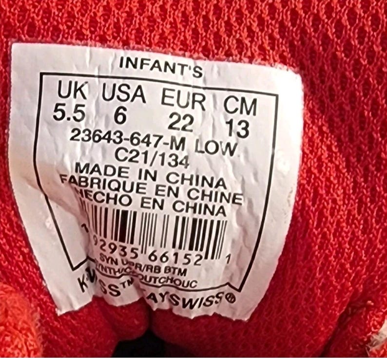 May include: Close-up of a label from a red infant's shoe. The label includes size information: UK 5.5, USA 6, EUR 22, and CM 13. The label also includes the text "MADE IN CHINA" and a barcode.