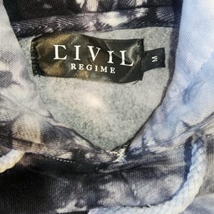 May include: A tie-dye hoodie in shades of black, white, and light blue. The label reads "CIVIL REGIME" in white text on a black background. The hoodie has a drawstring and a medium size tag.