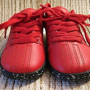May include: A pair of bright red sneakers with matching laces. The shoes have a stitched design on the toe and a black sole speckled with white. The sneakers are likely for a child.