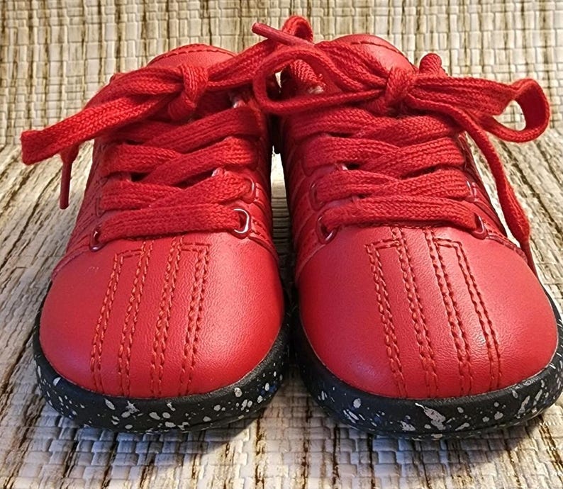 May include: A pair of bright red sneakers with matching laces. The shoes have a stitched design on the toe and a black sole speckled with white. The sneakers are likely for a child.