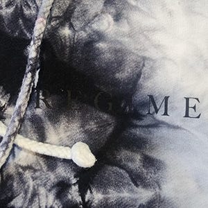 May include: A tie-dye hoodie in shades of black and white. The drawstring is white, and the word "GAME" is visible on the fabric. The hoodie has a classic design.