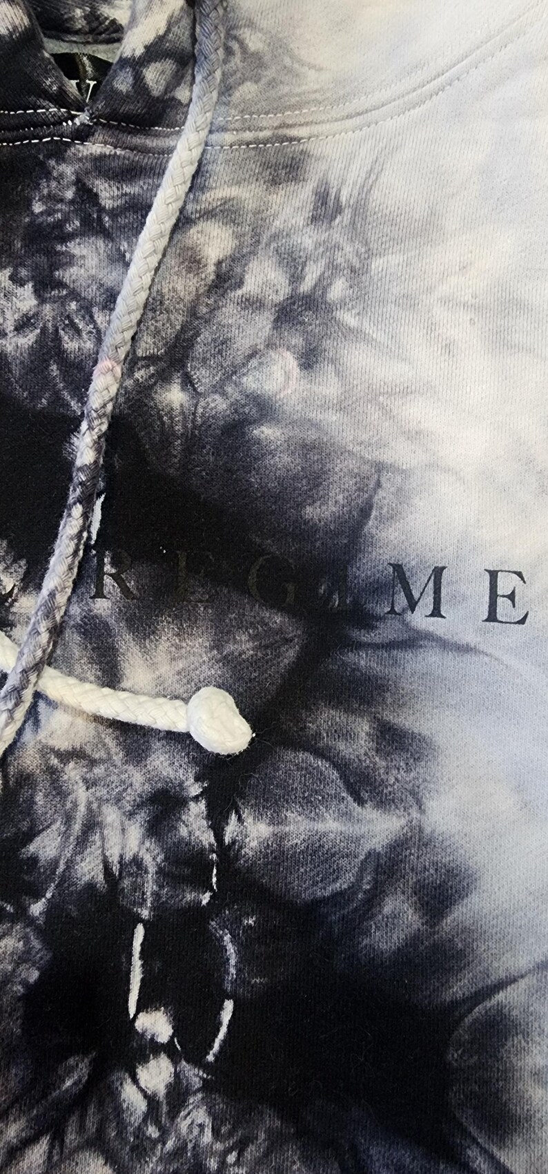 May include: A tie-dye hoodie in shades of black and white. The drawstring is white, and the word "GAME" is visible on the fabric. The hoodie has a classic design.