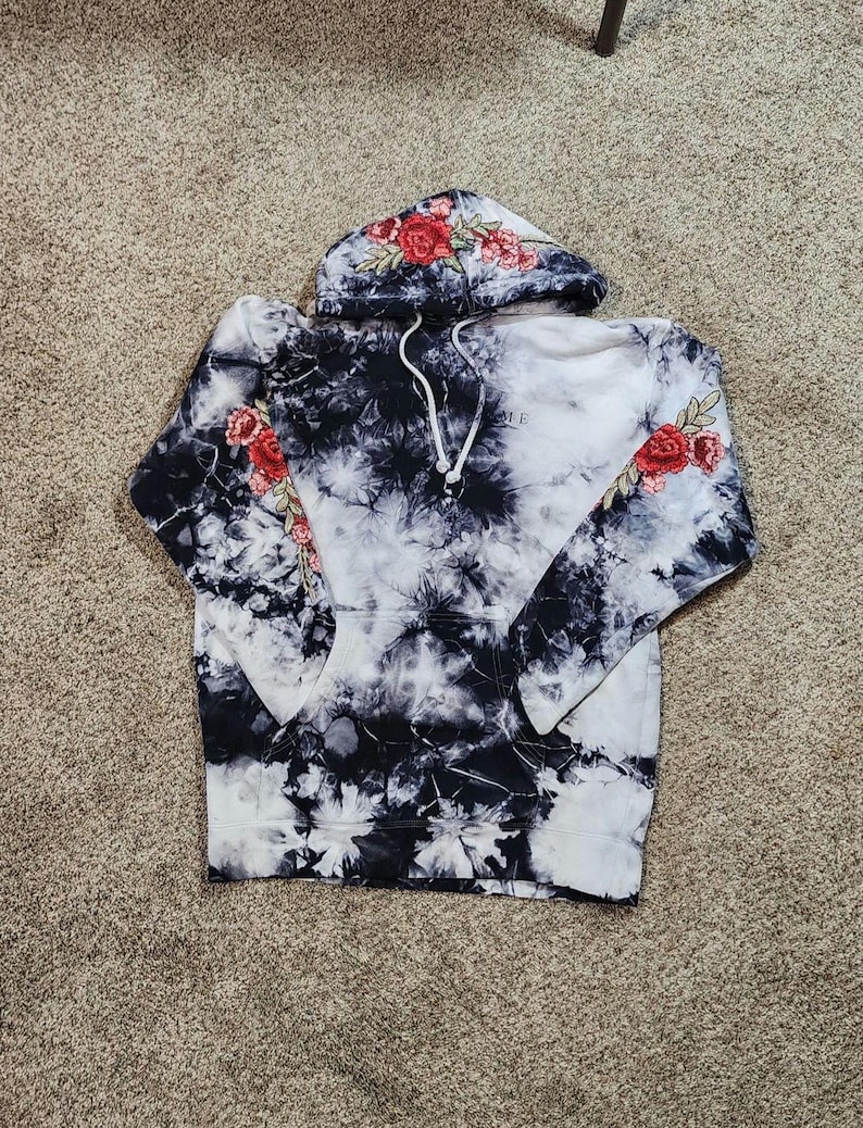May include: A tie-dye hoodie in black and white with red rose embroidery on the hood and sleeves. The word "ME" is printed on the front. The hoodie has a drawstring and a kangaroo pocket.